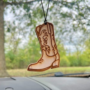 May include: A wooden cowboy boot car air freshener hanging from a rearview mirror. The boot is light brown and has a detailed design.
