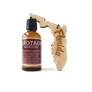 May include: A brown glass bottle with a black cap and a label that reads "AROTAGS MADE IN USA Beach Bum Fragrance Oil" and instructions for use. The bottle is next to a wooden cutout of the state of Florida.