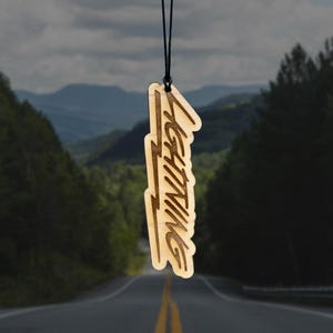 Puede incluir: Un ambientador de madera con la palabra "LIGHTNING" en una fuente estilizada. El ambientador es de color marrón claro y cuelga de una cuerda negra. El fondo muestra una carretera que serpentea por un paisaje montañoso.