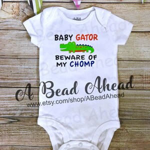 May include: White baby onesie with a green alligator graphic and the text "Baby Gator Beware of My Chomp". The onesie has a snap closure and is made of a soft, comfortable material.