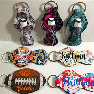 May include: Seven different lip balm holders with keychains. The holders are made of neoprene and have various patterns including paisley, butterflies, camouflage, peacocks, elephants, tie dye, and a football with the words 'UF Gators'.