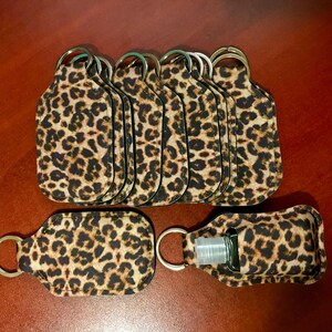 May include: A collection of leopard print hand sanitizer holders with key rings. The holders are brown and black with a repeating spotted pattern. One holder contains a clear bottle of hand sanitizer.