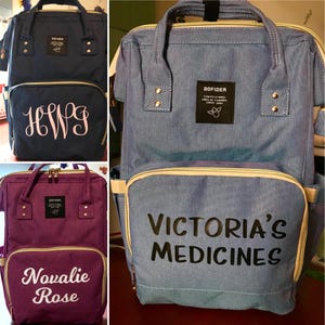 May include: Three different diaper bags in navy blue, denim blue, and purple. Each bag has a front pocket with custom text in white. The navy bag has the initials "HWG", the denim bag says "Victoria's Medicines", and the purple bag says "Novalie Rose".