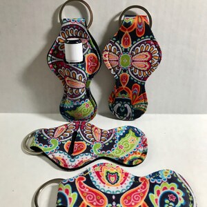 May include: Four colorful keychains with a paisley pattern. Each keychain has a small pocket for holding a lip balm or essential oil roller. The keychains are made of fabric and have a silver metal ring.