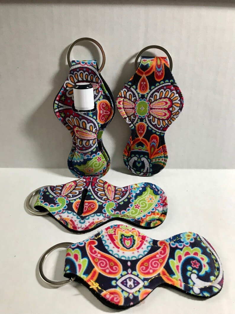 May include: Four colorful keychains with a paisley pattern. Each keychain has a small pocket for holding a lip balm or essential oil roller. The keychains are made of fabric and have a silver metal ring.