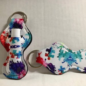 May include: Two white fabric keychains with a colorful paint splatter and puzzle piece design. One keychain has a small clear tube for lip balm or hand sanitizer. Both keychains have a silver metal ring.