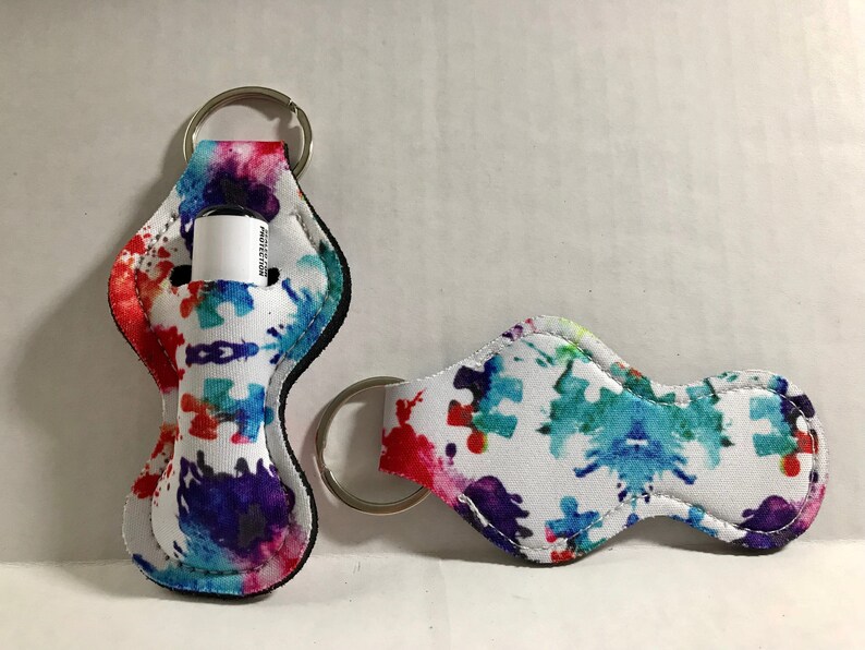 May include: Two white fabric keychains with a colorful paint splatter and puzzle piece design. One keychain has a small clear tube for lip balm or hand sanitizer. Both keychains have a silver metal ring.