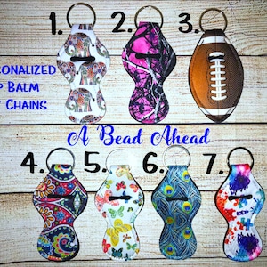 May include: Seven different lip balm keychains with various designs. The designs include elephants, camouflage, a football, paisley, butterflies, peacocks, and tie-dye. The keychains are labeled with numbers 1 through 7. The text "Personalized Lip Balm Key Chains" and "A Bead Ahead" are also visible.