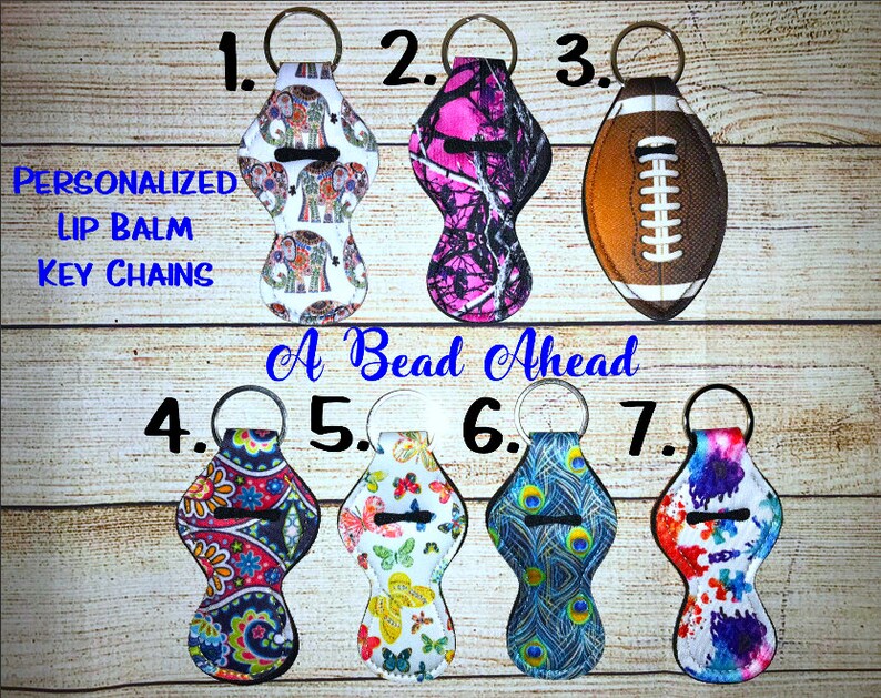 May include: Seven different lip balm keychains with various designs. The designs include elephants, camouflage, a football, paisley, butterflies, peacocks, and tie-dye. The keychains are labeled with numbers 1 through 7. The text "Personalized Lip Balm Key Chains" and "A Bead Ahead" are also visible.
