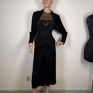 May include: A black dress with a matching bolero jacket displayed on a mannequin. The dress features a sheer neckline and lace detailing on the bodice. The jacket has long sleeves. The mannequin is positioned in front of a plain white wall.