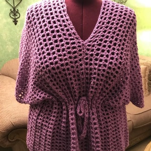 May include: A lavender crochet top with a V-neck and short sleeves. The top has a drawstring waist and a loose, open weave design. The garment is displayed on a maroon dress form.