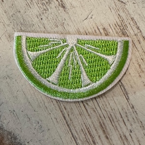 May include: Embroidered patch in the shape of a lime slice. The patch features a bright green interior with white detailing and a white border. The design is a detailed representation of a lime.