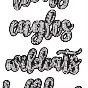 May include: Four iron-on patches with team names: "Lions", "Eagles", "Wildcats", and "Bulldogs". Each word is in a cursive font, outlined in black, and filled with silver sequins. The background is white.