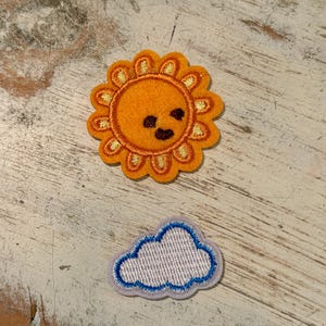 May include: An embroidered patch featuring a bright orange sun with a smiling face and golden yellow rays, positioned above a smaller embroidered patch of a light purple cloud with a white center and a blue stitched outline border.