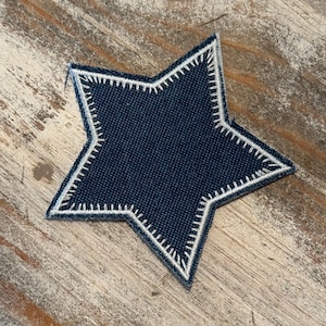 May include: A navy blue star-shaped patch with white stitching around the edges. The patch is made of a textured fabric and is set against a light brown wooden surface.