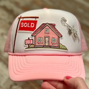 May include: Pink and white trucker hat with a pink brim. The hat features a patch of a pink house with the word "SOLD" on a sign. A silver charm of a house and keys is pinned to the hat.