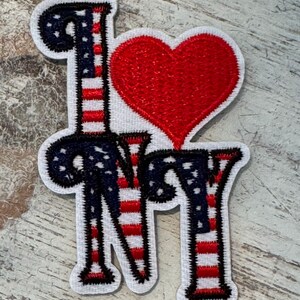 Pu&ograve; includere: Una toppa ricamata con le parole "I Love NY" in un font stilizzato. Le lettere sono contornate di nero e riempite con strisce rosse e bianche, che ricordano la bandiera americana. Un cuore rosso &egrave; posizionato tra la "I" e la "NY".