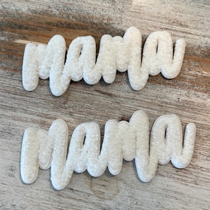 May include: Two white, fluffy "mama" word cutouts in a cursive font. The cutouts are made of a soft, textured material and are placed on a weathered, light brown wooden surface. The letters are slightly raised, creating a 3D effect.