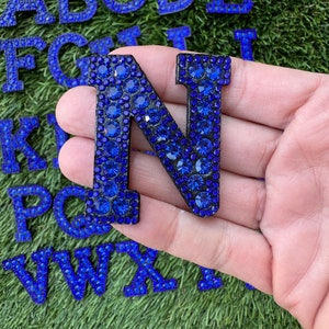 May include: A blue rhinestone letter N, with a black backing, is held in a hand. The letter is surrounded by other letters of the alphabet, also covered in blue rhinestones.