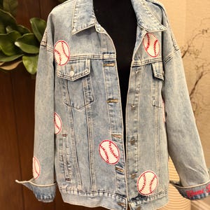May include: Light blue denim jacket featuring multiple embroidered baseball patches. The jacket has a classic design with button closures, pockets, and a collared neckline. The cuffs are rolled up, revealing red embroidered text. A stylish and sporty outerwear piece.