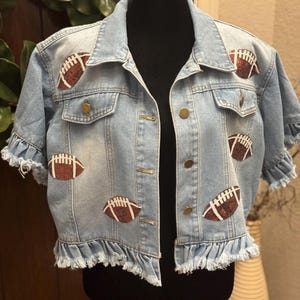 May include: Light blue denim jacket with short sleeves and a ruffled hem. The jacket is adorned with multiple brown and white football appliqués. Features brass-colored buttons and a classic collar. A stylish, sporty fashion item.