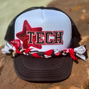 May include: Black and white trucker hat with a white front panel featuring a red star and the word "TECH" in red and black lettering. The hat has a black mesh back and a braided band with red and white accents.