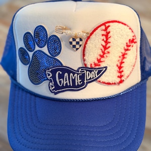 May include: A blue and white trucker hat with a baseball theme. The hat features a sequined paw print, a baseball, and a banner that reads "GAME DAY." A decorative pin with a heart is also attached.