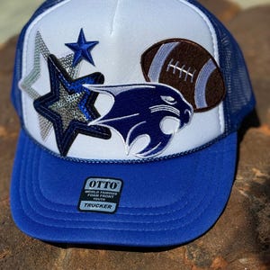 May include: White and blue trucker hat with a blue panther, a brown football, and a silver star with sequins. The hat has a blue mesh back and a black label that says "OTTO WORLD FAMOUS FOAM FRONT YOUTH TRUCKER".