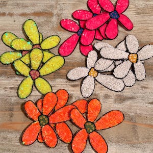 May include: A collection of sequined flower appliques in various colors, including yellow, pink, white, and orange. Each flower has a contrasting center and black outline, perfect for embellishing clothing or accessories.