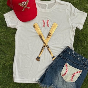 May include: A white t-shirt with a gold sequin baseball bat and ball design. A red mesh baseball cap with a white baseball bat and ball design. Blue denim shorts with a white baseball pocket with red stitching.