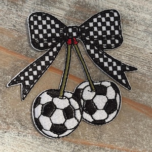 May include: Embroidered patch featuring two cherries with soccer ball designs, connected by green stems and a black and white checkered bow. The patch has a white border and is set against a neutral background.
