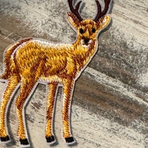 May include: A brown embroidered deer patch with white outlining. The deer has large antlers and is standing on all four legs.