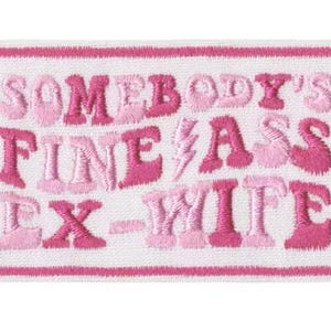 May include: Embroidered patch with a white background and a pink border. The patch features the text "SOMEBODY'S FINE ASS EX-WIFE" in shades of pink. The text is in a playful, stylized font.