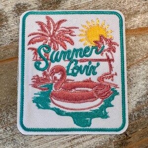 May include: Embroidered patch with a white background, featuring a flamingo float, palm trees, and a yellow sun. The words "Summer Lovin'" are written in teal script. The patch has a teal border.
