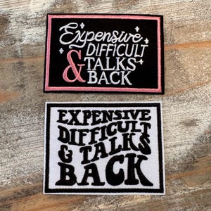 May include: Two rectangular patches with the text "Expensive Difficult Talks Back". One patch is black with pink accents, the other is white with black text. The patches are on a wooden surface.