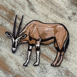 May include: Embroidered patch of a brown and white oryx, a type of antelope, standing on a light brown wooden surface.