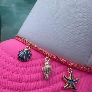 May include: A pink trucker hat with a white front panel and a gold chain detail. The hat features three charms: a blue seashell, a silver conch shell, and a blue and gold starfish. The hat is a summer accessory.