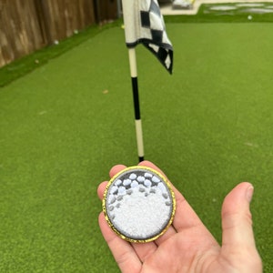 May include: A white golf ball patch with a gold border. The patch is embroidered with a realistic golf ball design.
