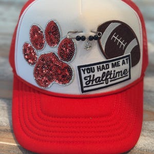 May include: Red and white trucker hat with a red brim and mesh back. The hat features a sequined paw print, a football patch, and a patch that reads "You Had Me at Halftime."