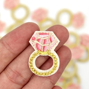 May include: A pink and gold embroidered ring patch with a large diamond design. The patch is on a white background.