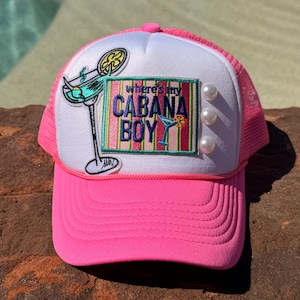 May include: A bright pink trucker hat with a white front panel featuring an embroidered patch that reads "where's my CABANA BOY" in blue and purple text. The patch includes a cocktail glass graphic and three white faux pearl embellishments.