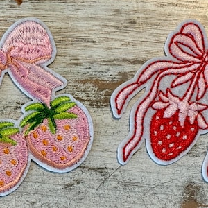 May include: Two embroidered iron-on patches featuring strawberries with pink bows. One patch has two strawberries and the other has one strawberry.