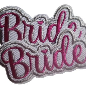 May include: Two white fabric patches with pink embroidered lettering that reads "Bride" in a cursive font.