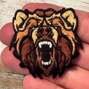 May include: Embroidered patch of a brown bear with a fierce expression, its mouth open in a growl. The bear's fur is detailed with brown and tan stitching.