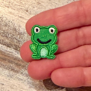 May include: A small, embroidered patch in the shape of a green frog. The frog has a smiling black mouth, white eyes with black pupils, and detailed stitching. The patch is likely for clothing or accessories.
