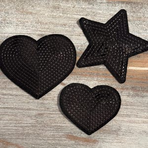 May include: Three black decorative shapes: a large heart, a star, and a smaller heart. Each shape is made of a woven material with a stitched design. The shapes are arranged on a light-colored wooden surface.