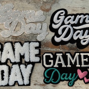 May include: Four different iron-on patches with the text "Game Day" in various styles. The patches are white with black outlines and feature different textures, including sequins, fluffy fabric, and embroidery.