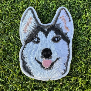 May include: Embroidered patch of a husky dog's face with black and white fur, a pink tongue sticking out, and a white background.