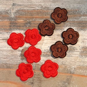 May include: A collection of embroidered flower patches in two colors. Six red flower patches with white borders are arranged with four brown flower patches. The patches are on a light brown wooden surface.
