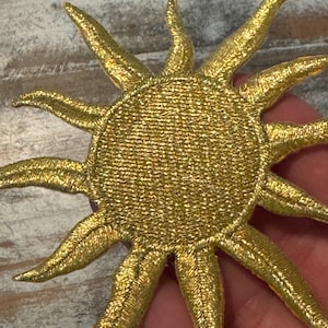 May include: A gold sun-shaped embroidered patch with a circular center and twelve radiating pointed rays. The surface of the patch features a dense, metallic-toned thread texture that creates a shimmering effect across the entire design.